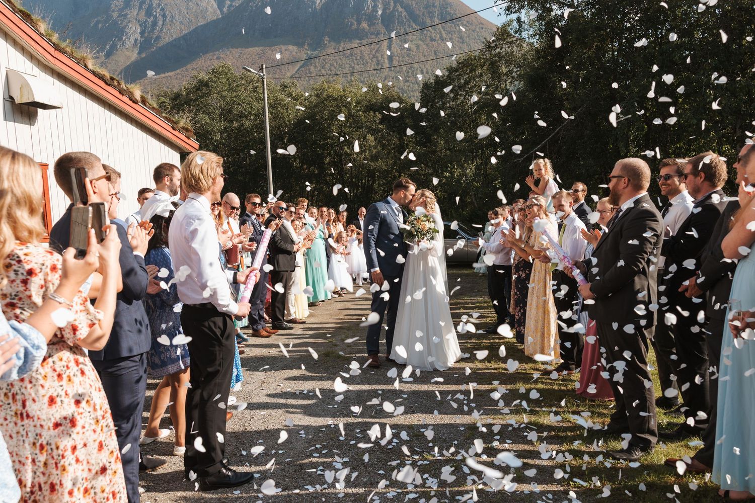 Wedding guests toss white confetti as couple celebrates at outdoor ceremony with stunning mountain backdrop.