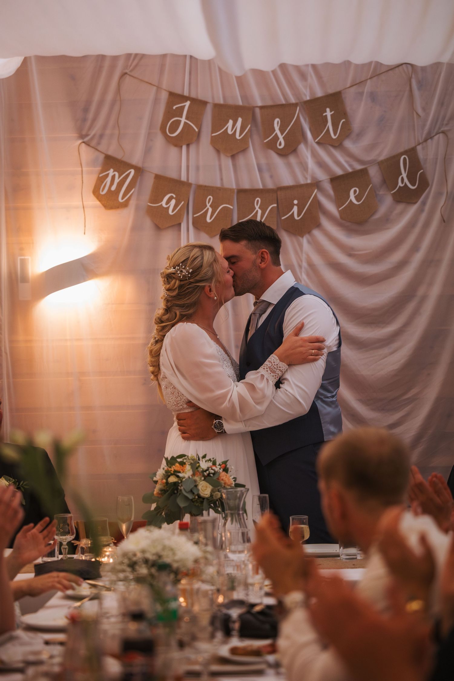 A romantic wedding reception scene with a 'Just Married' banner, floral centerpieces and guests clapping at a candlelit venue.
