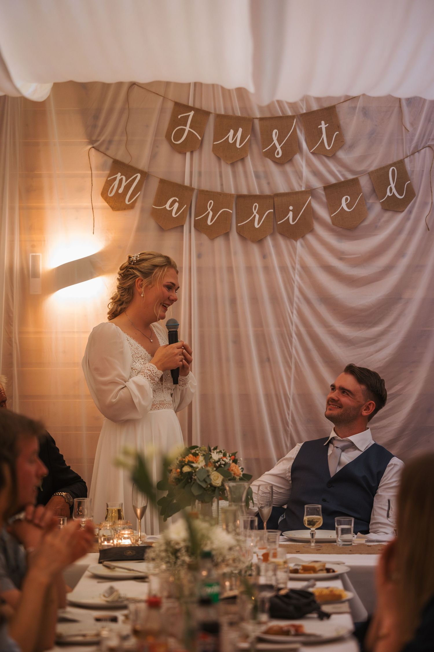 A wedding reception with 'Just Married' banner hanging above, showing couple at head table during evening speeches.