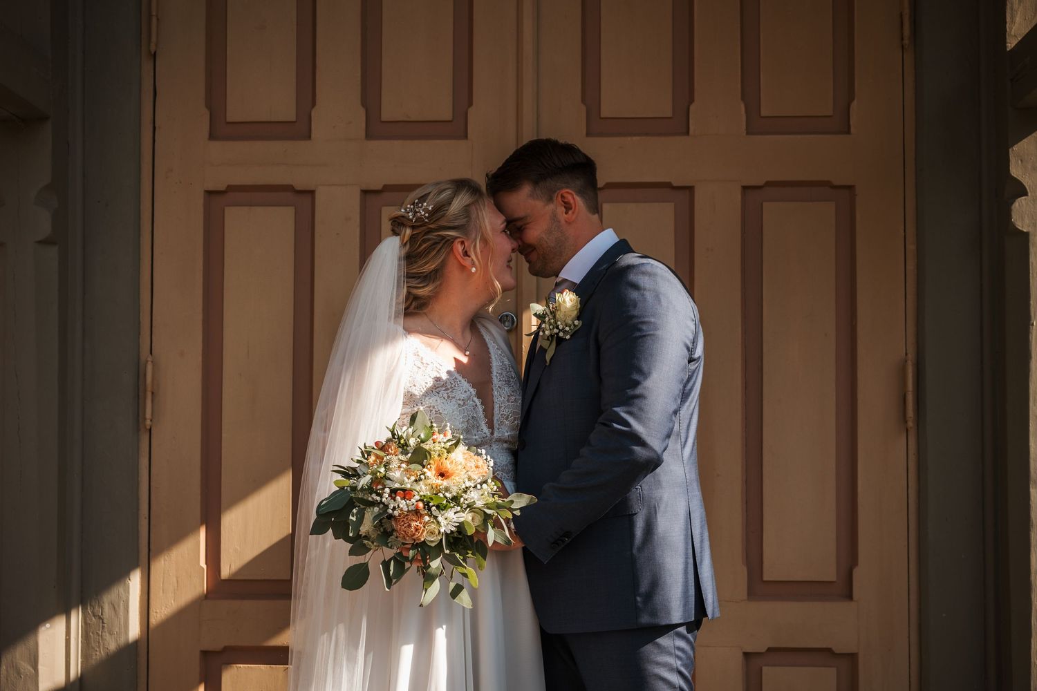A romantic wedding couple shares an intimate moment in front of a wooden door while holding a colorful bouquet.