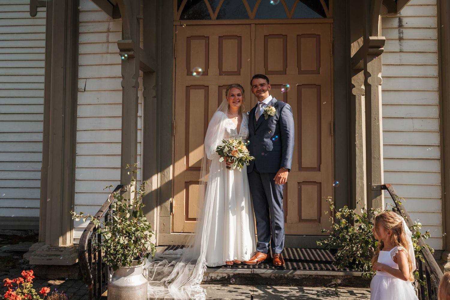 Wedding party poses for photos on the front porch of a classic white house with wooden columns.