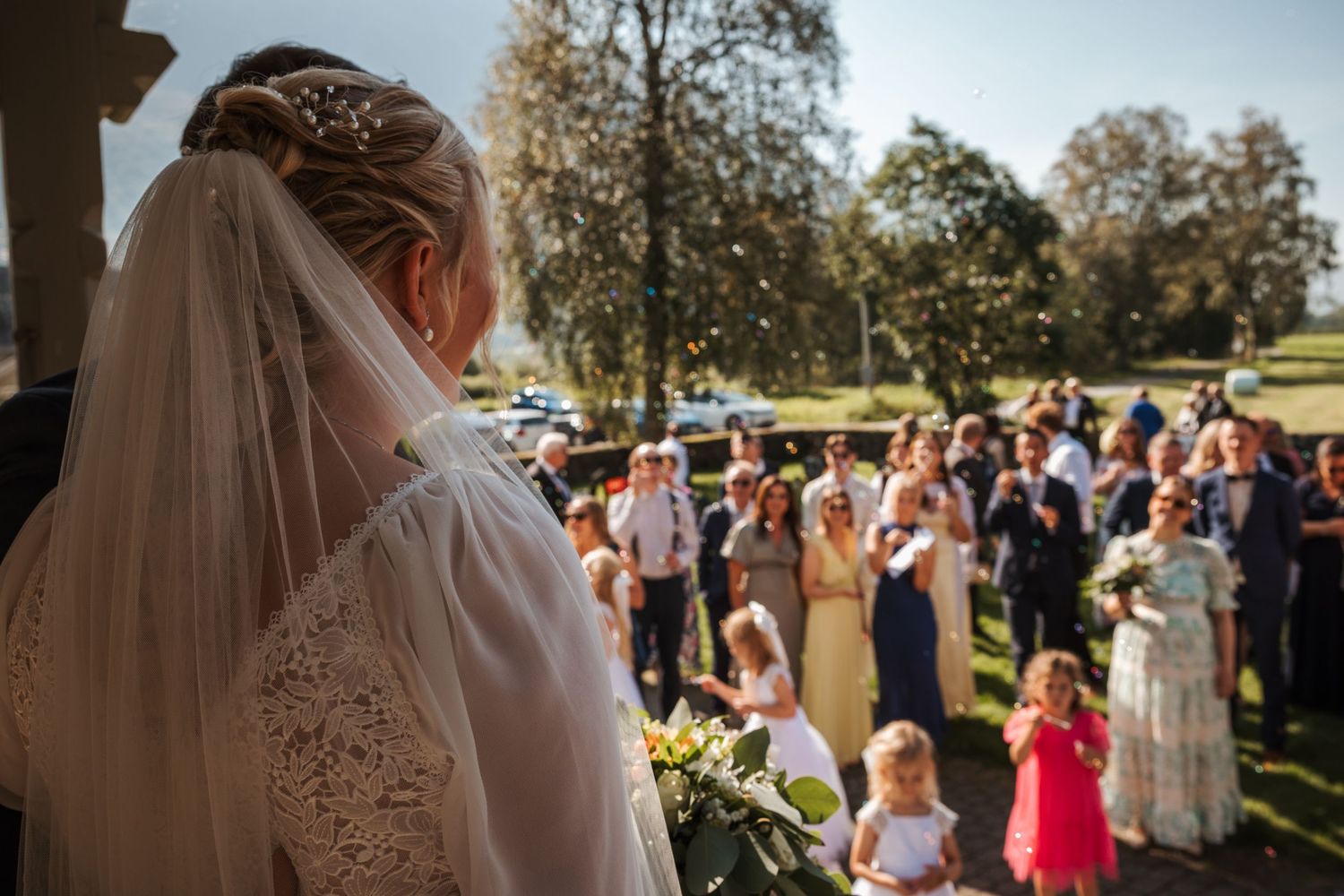 Wedding guests gather on a sunny lawn during an outdoor ceremony as a bride stands at the front holding a bouquet.