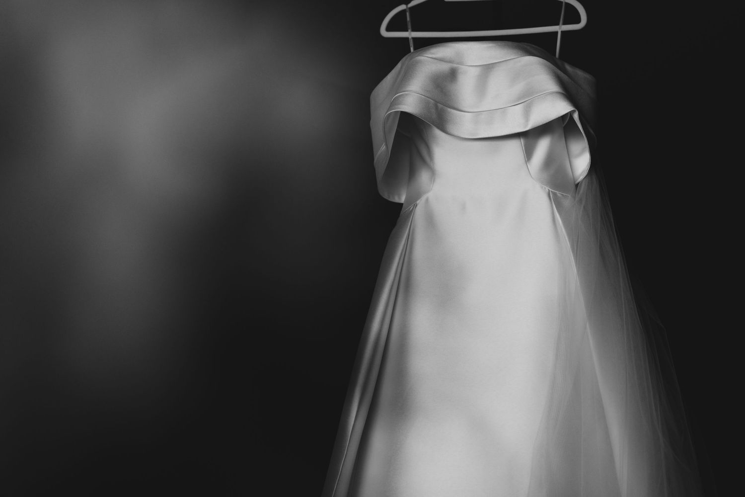 Elegant white wedding dress hangs gracefully against dark background in black and white.