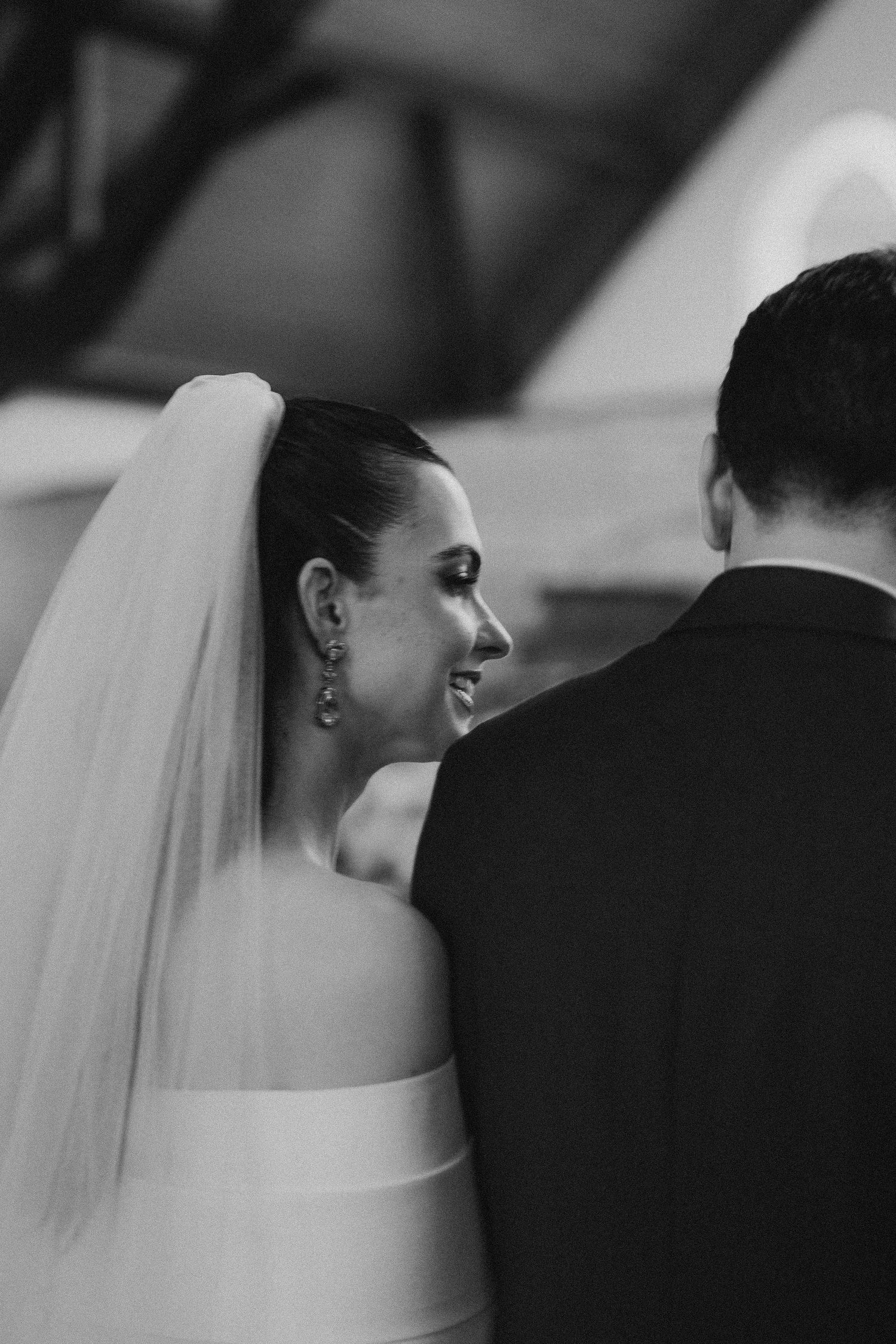 Black and white photo of a bride in veil glancing over her shoulder during the ceremony.