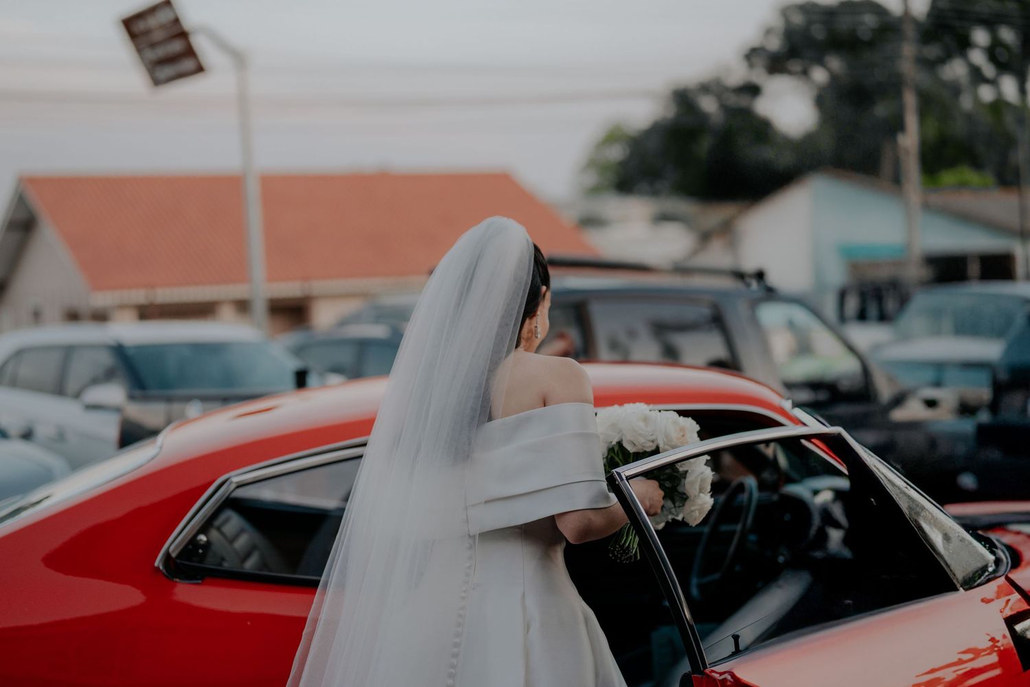 Bride in flowing white veil getting into a red car after the wedding ceremony.