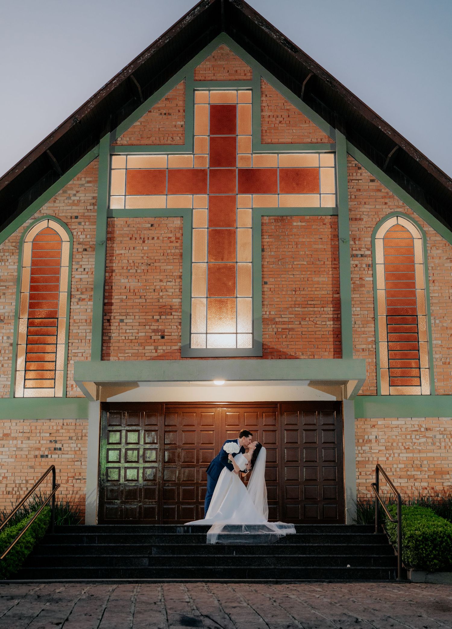 Newlyweds pose for romantic portrait in front of brick church with prominent cross design.