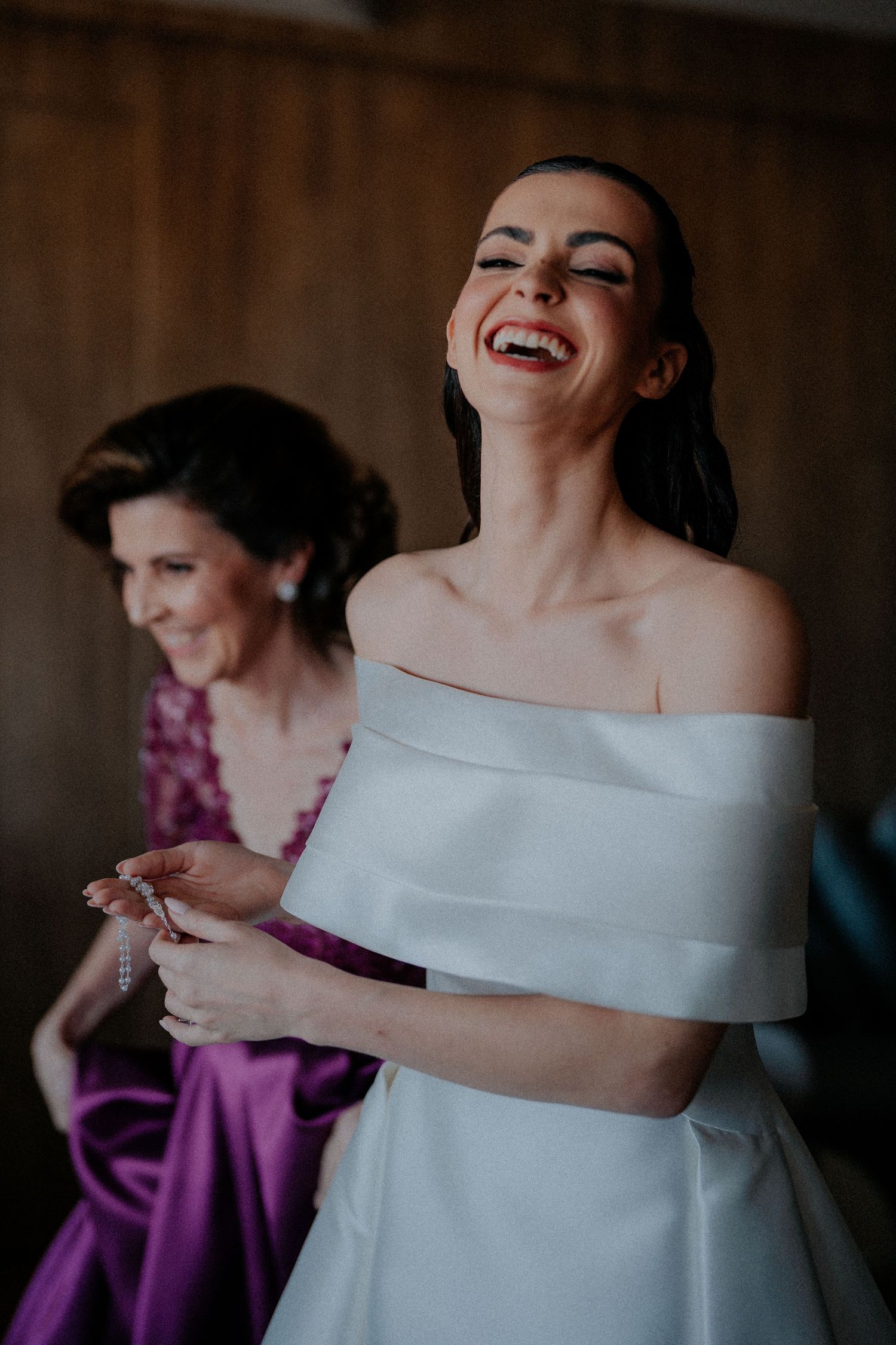 Bride in off-shoulder wedding dress laughs joyfully during pre-wedding preparations.