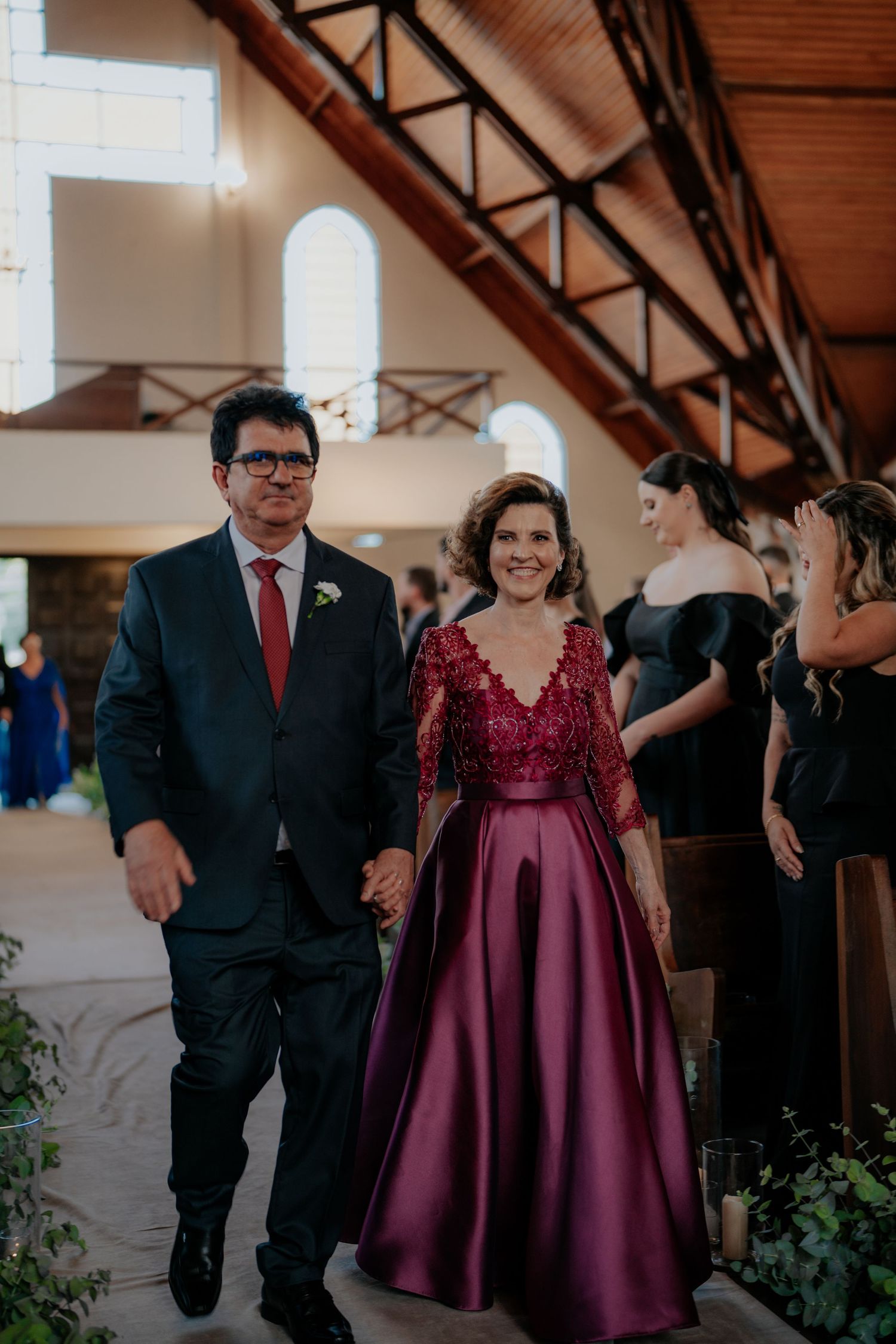 Wedding party members walk and dance down indoor venue aisle decorated with greenery.