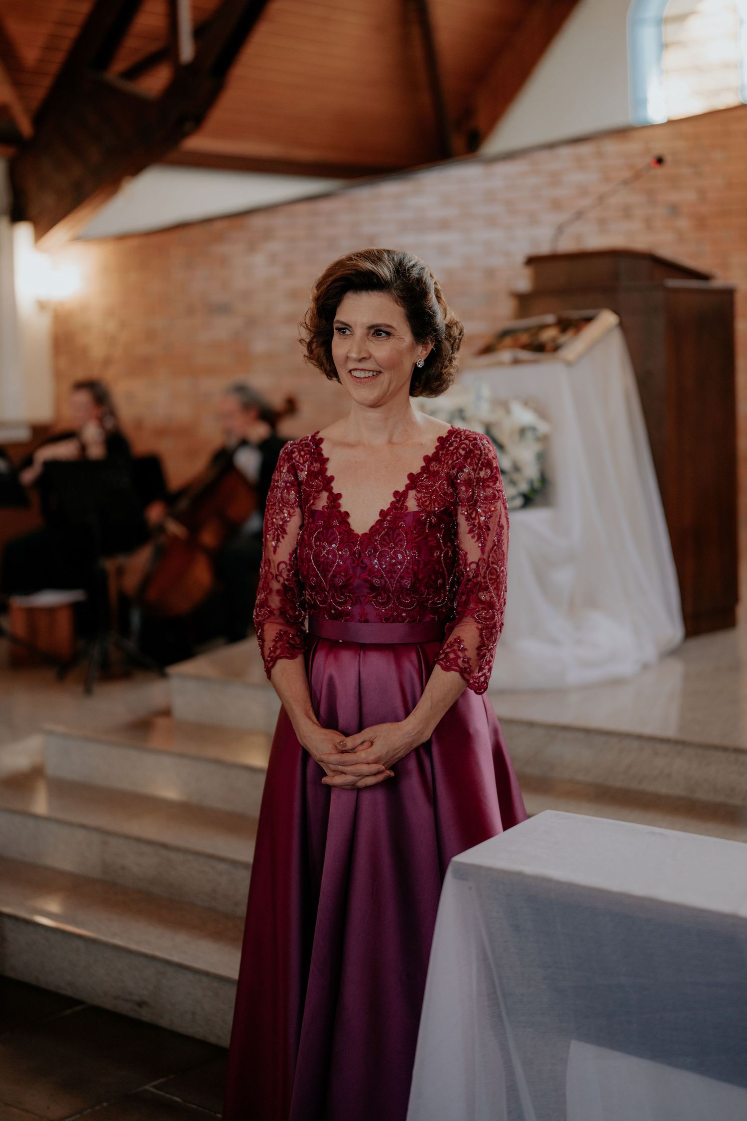Woman in burgundy lace dress stands elegantly in indoor wedding venue.