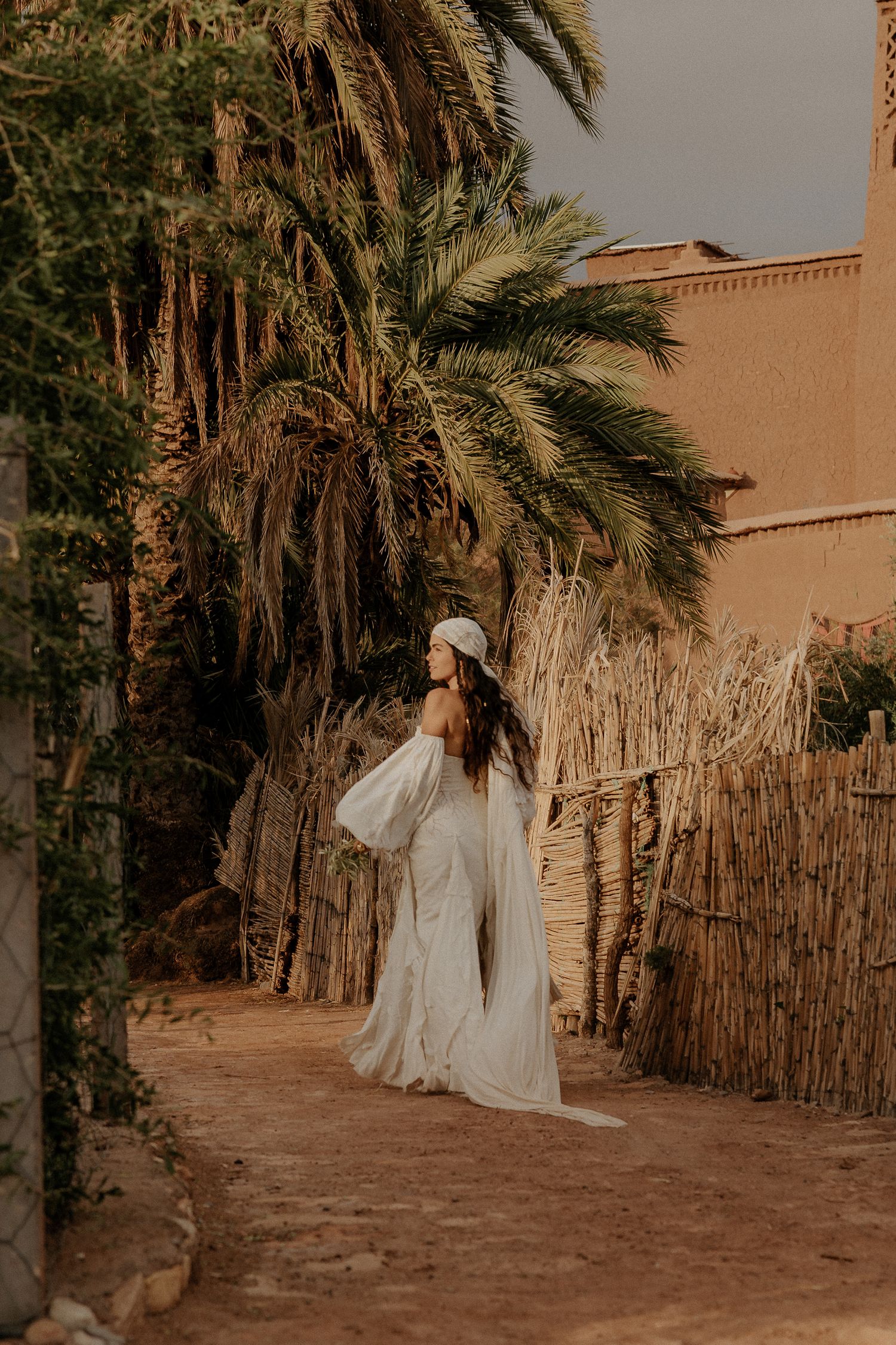 White dress flows elegantly along palm-lined pathway in desert setting.