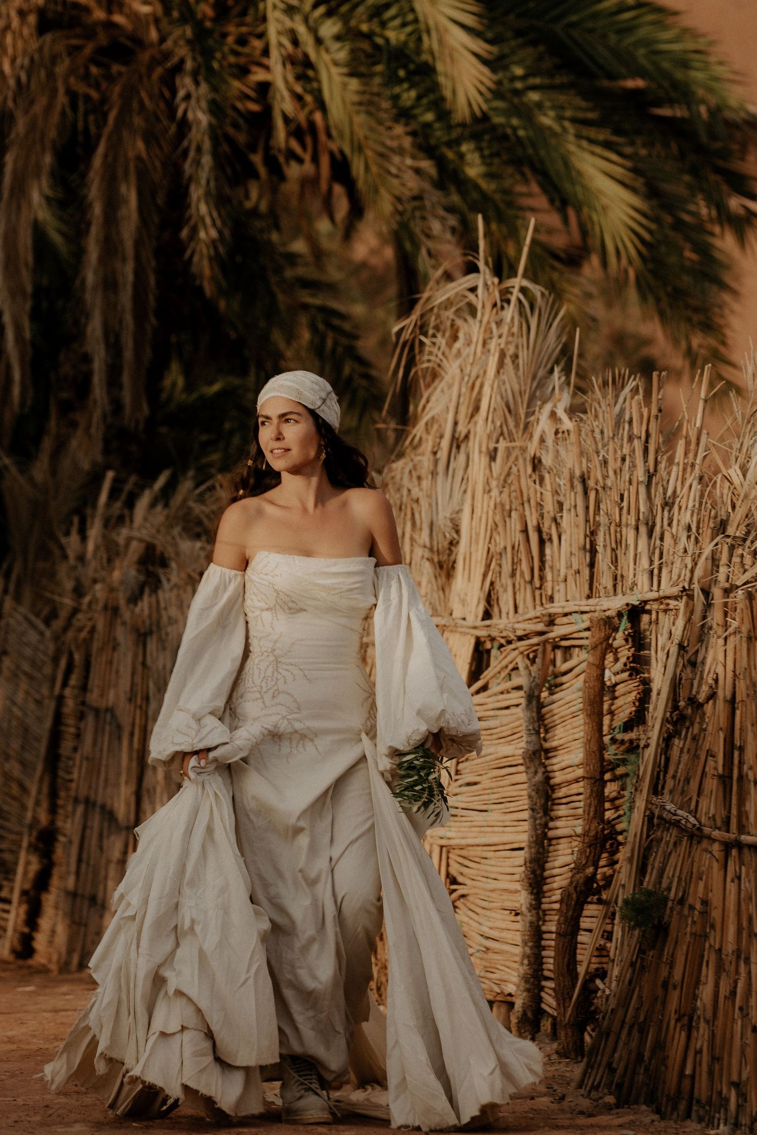A flowing white dress contrasts against palm trees and reed fencing in a desert setting at sunset.