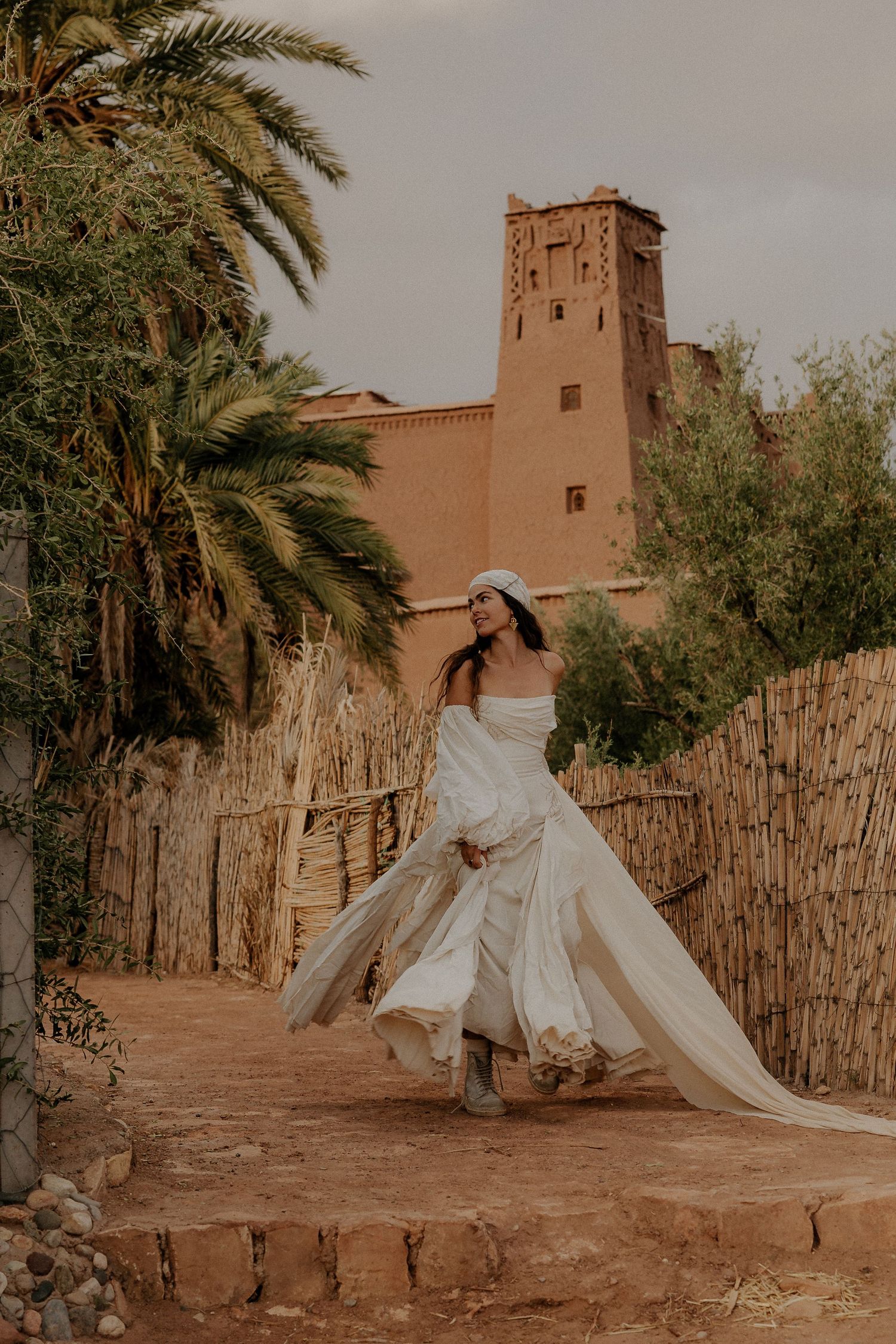 White flowing dress billows dramatically against ancient desert architecture and palm trees.