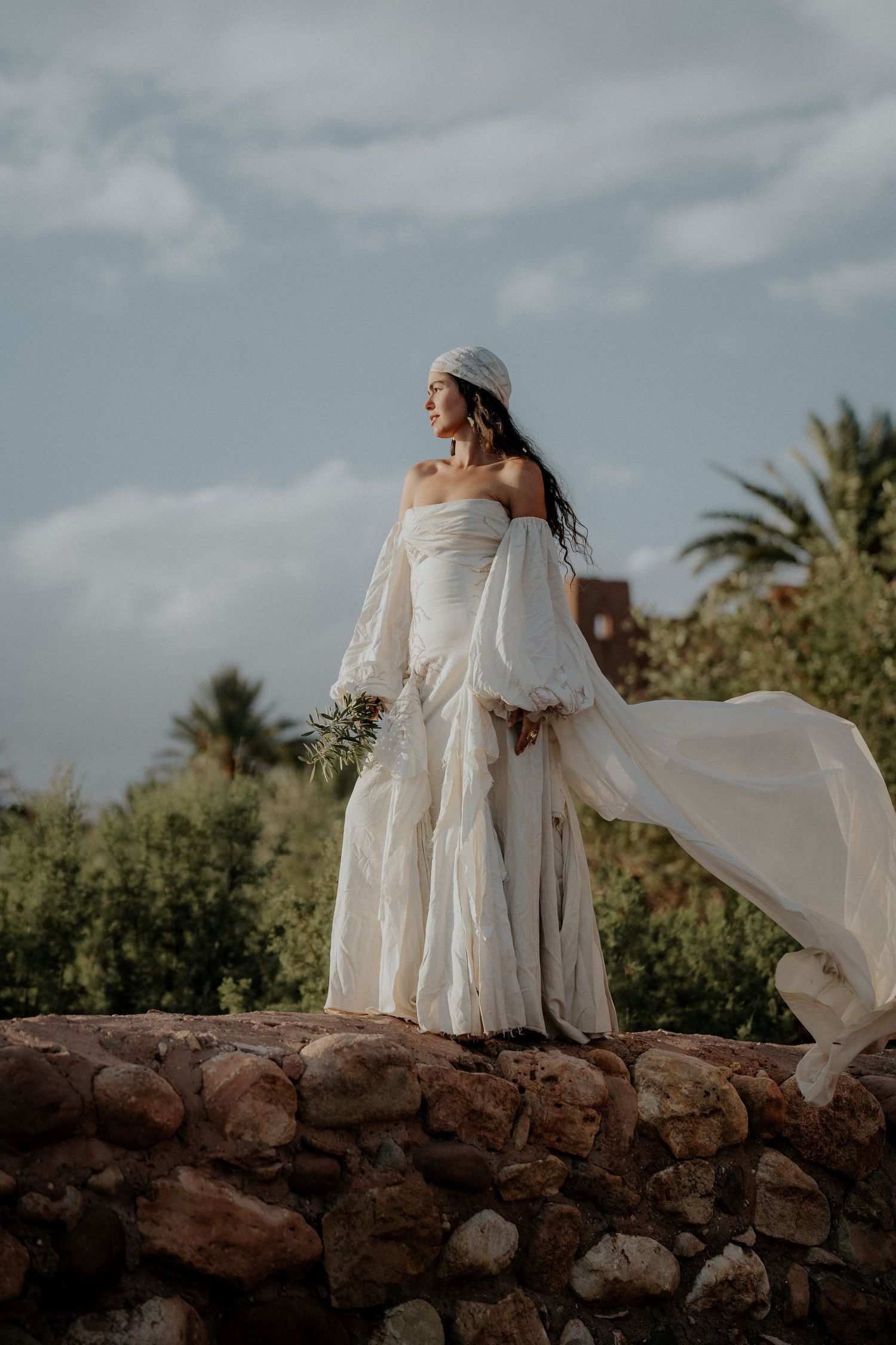 Figure in white dress stands gracefully on stone wall with palm trees in background.