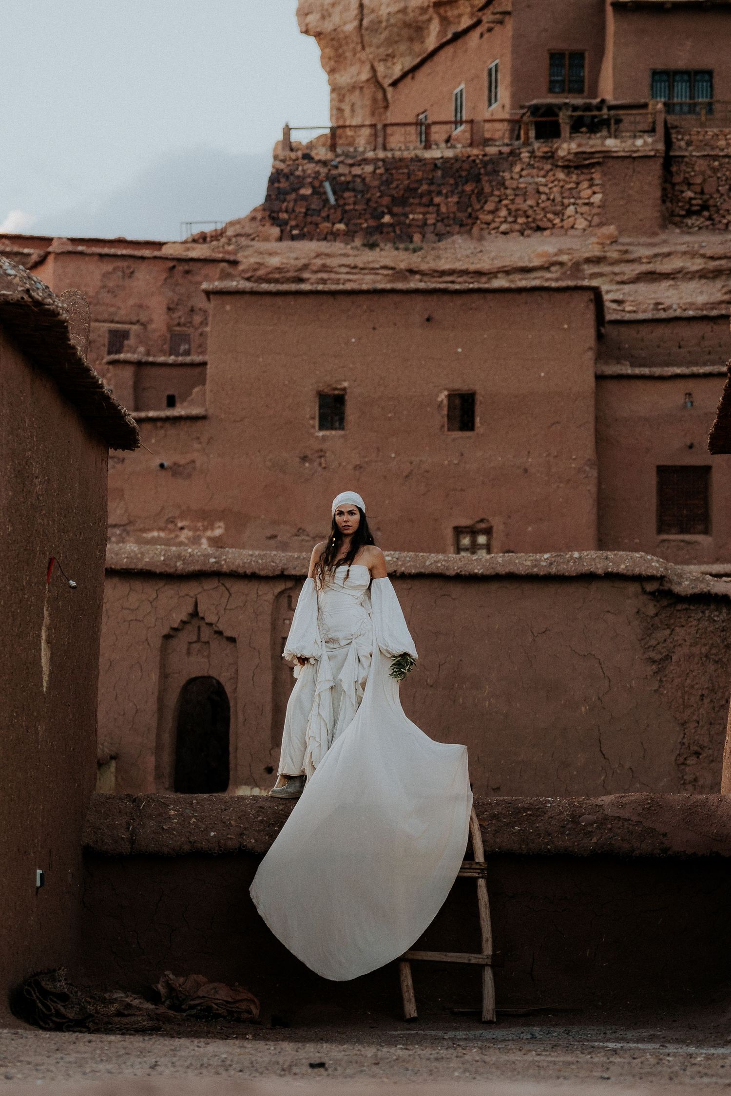 White dress creates dramatic contrast against traditional clay buildings in desert setting.