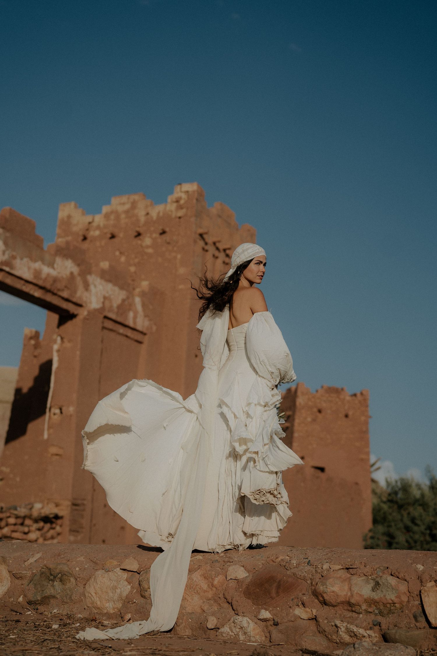 Flowing white dress catches the wind against adobe walls in a desert setting.