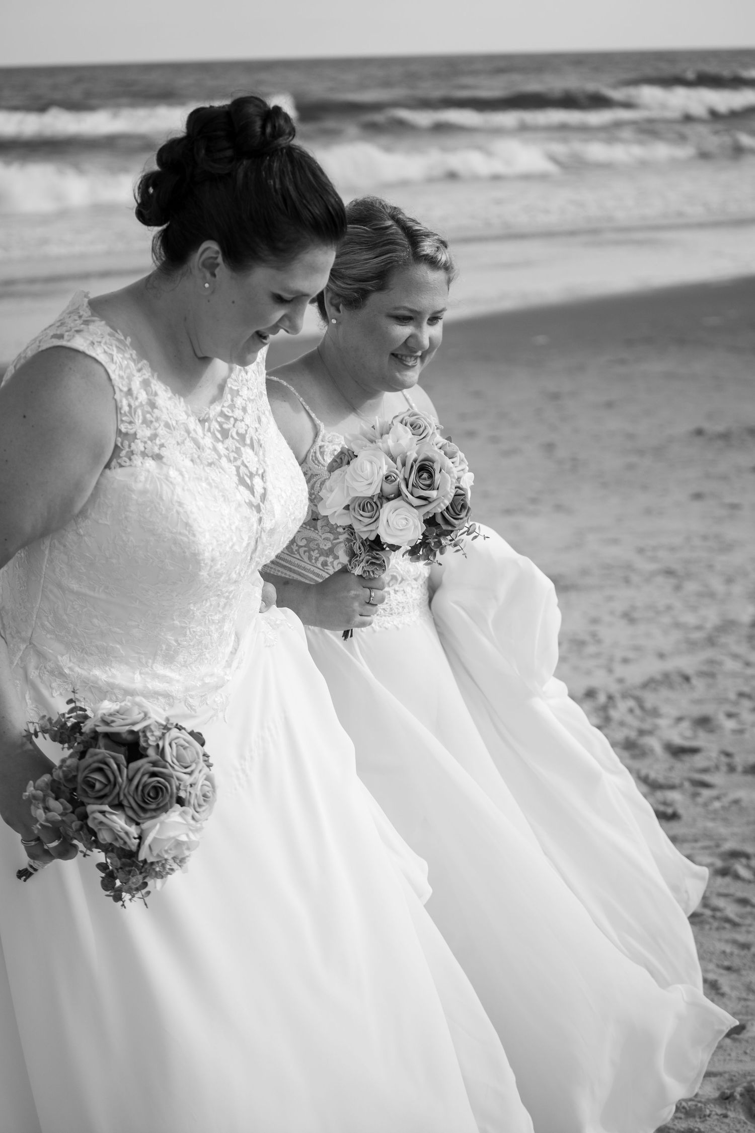 Two brides in white wedding dresses holding bouquets while walking on a beach.