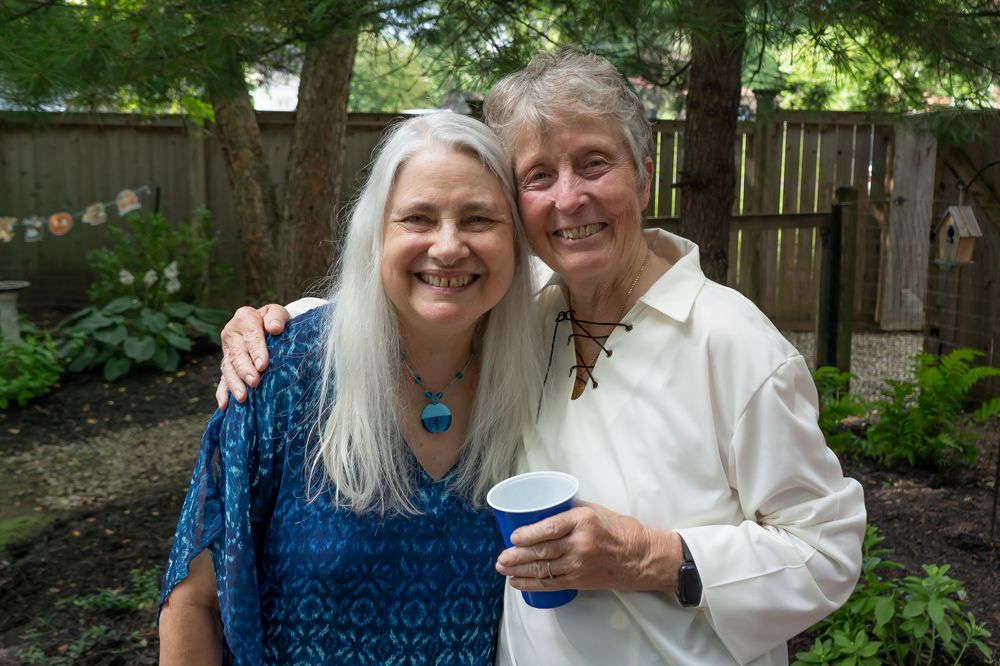 Two people enjoy a picnic outdoors near trees while celebrating with beverages on a sunny day.