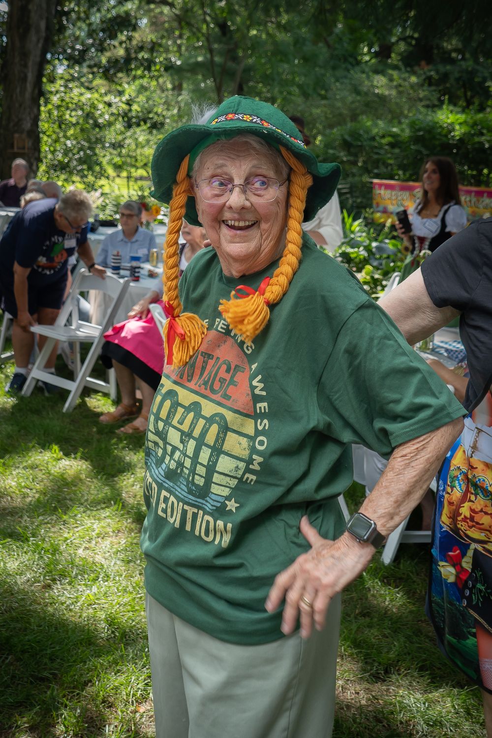Person in a green t-shirt smiles and gestures energetically at an outdoor summer gathering.