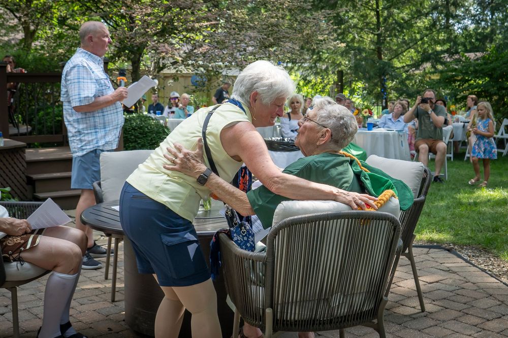 People gather for an outdoor social event seated on patio furniture in a garden setting.