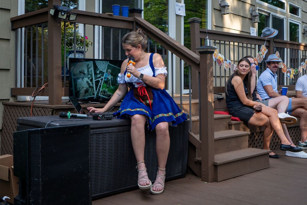 People in casual attire sit on wooden benches at a party on a sunny day.