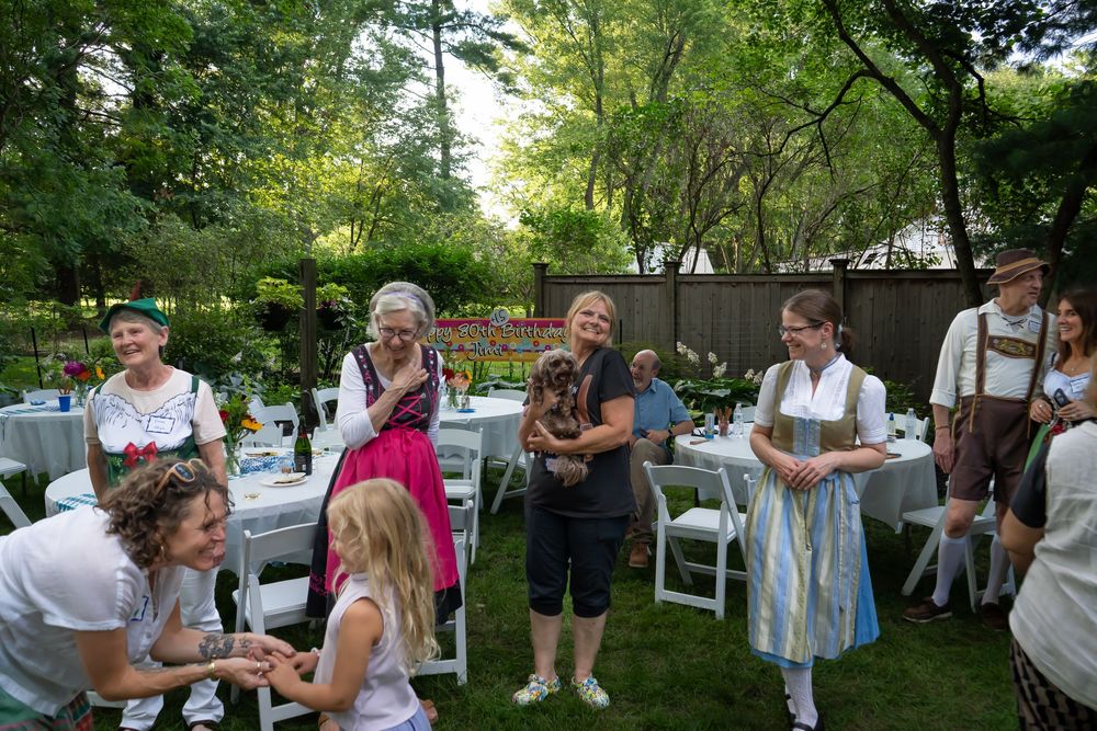 Group of people gathered outdoors at casual summer party in backyard setting with trees and greenery.
