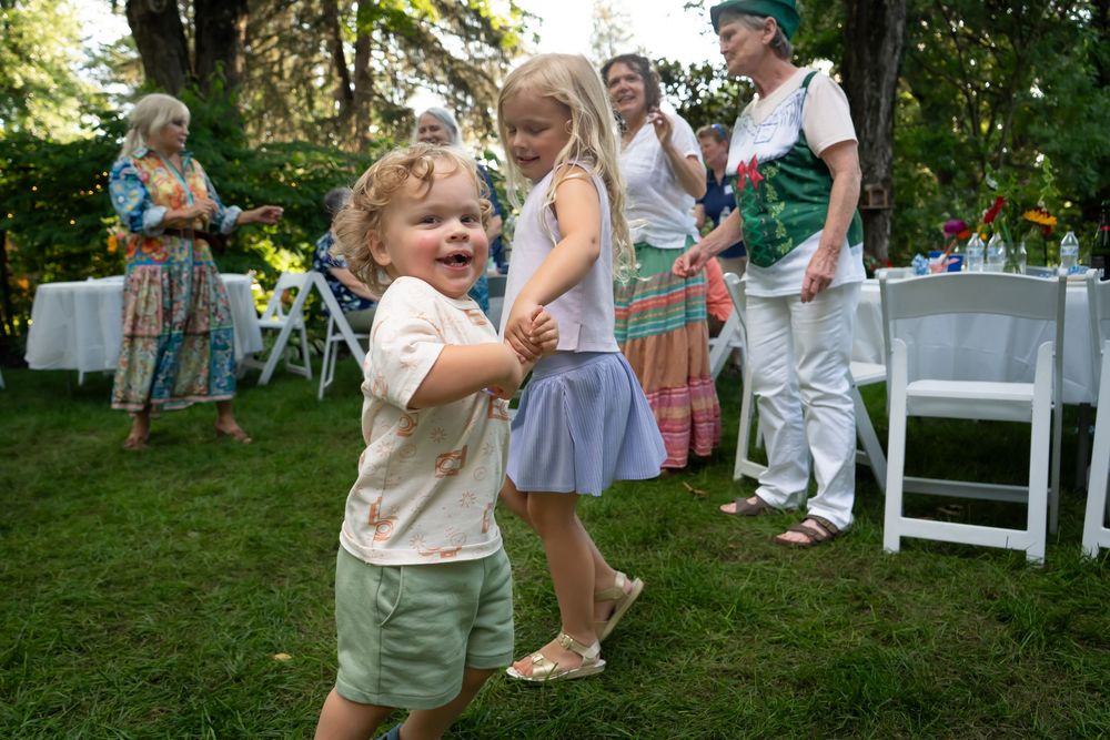 People gather outdoors for a festive celebration on a lawn with chairs and bright greenery in the background. children dancing.