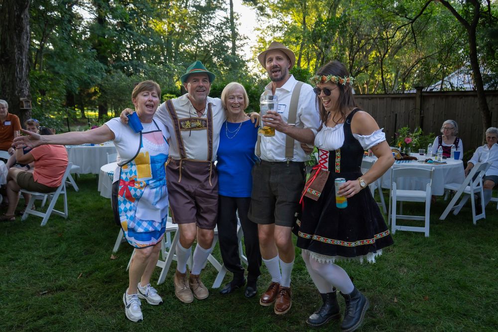 Groups of festive Oktoberfest attendees in traditional German dress gather outdoors at tables and chairs during celebrations.