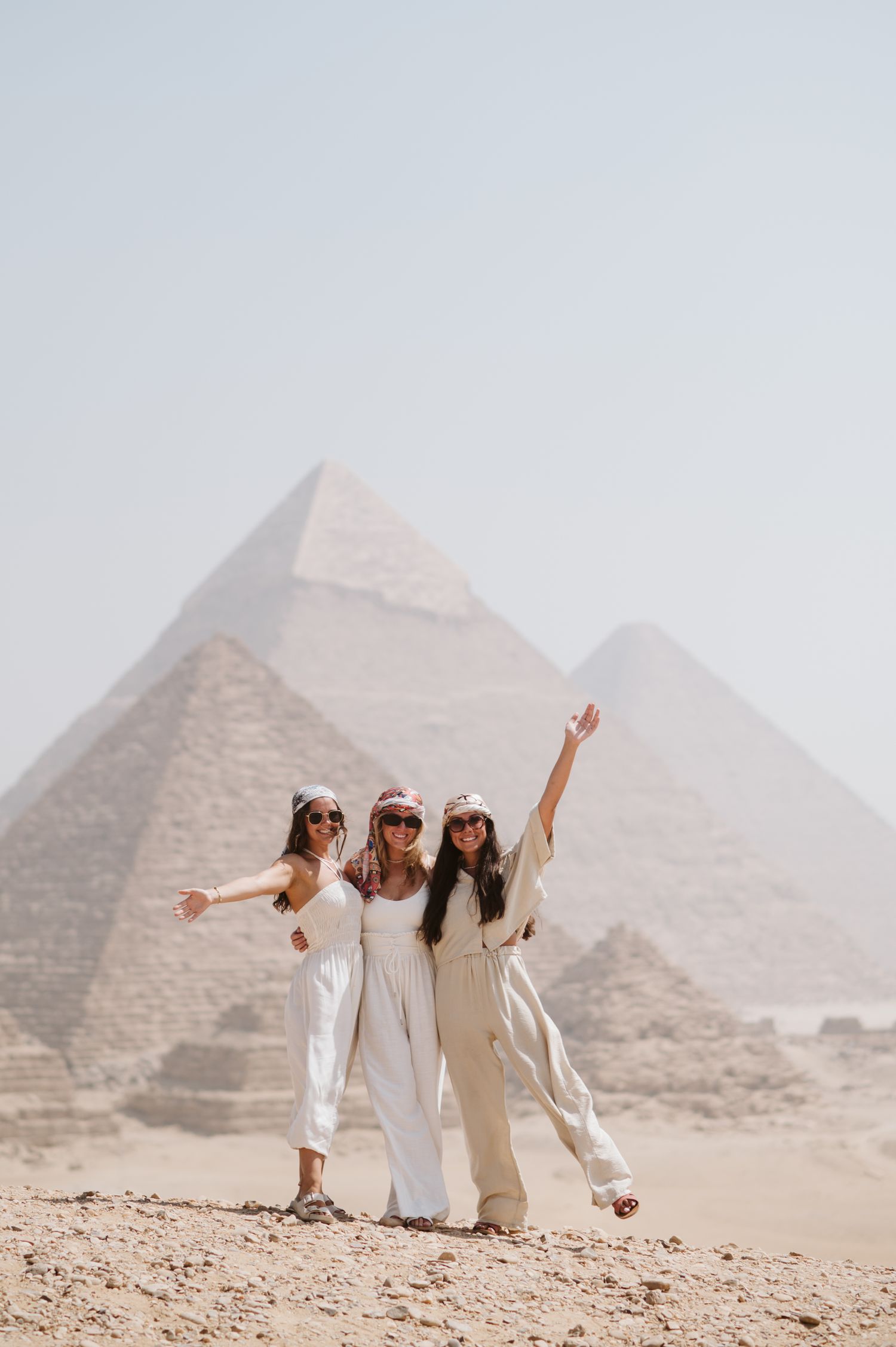 Three tourists in white outfits posing joyfully in front of the Egyptian pyramids.