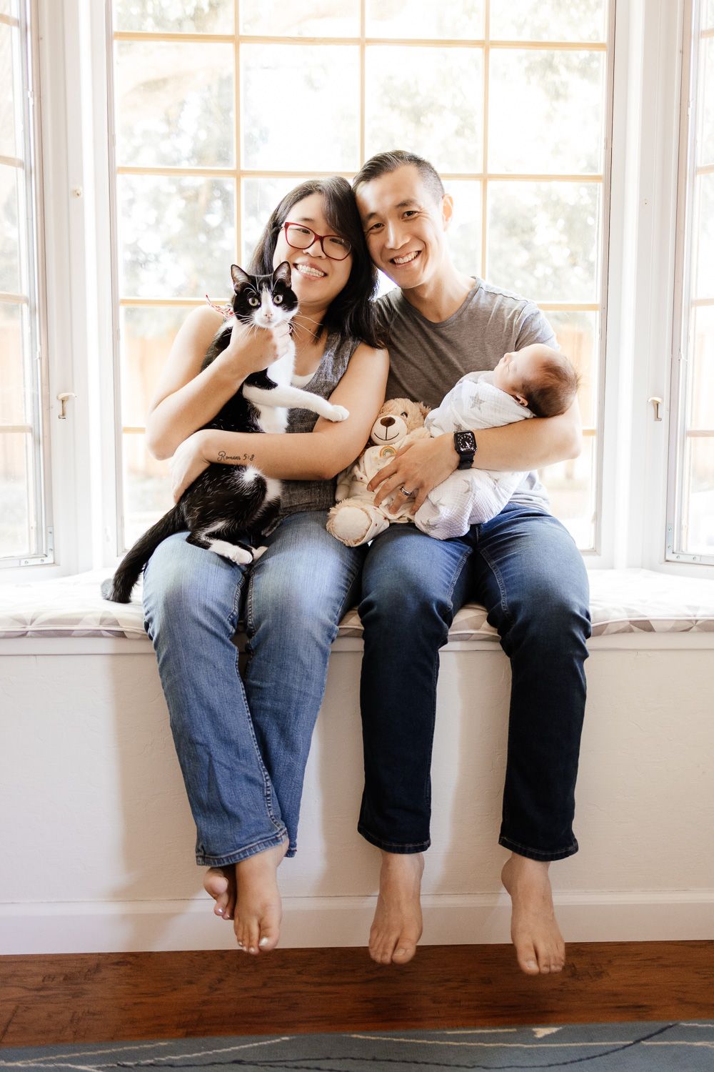 Parents smile while sitting in their airy and bright bay window with their newborn baby and cat
