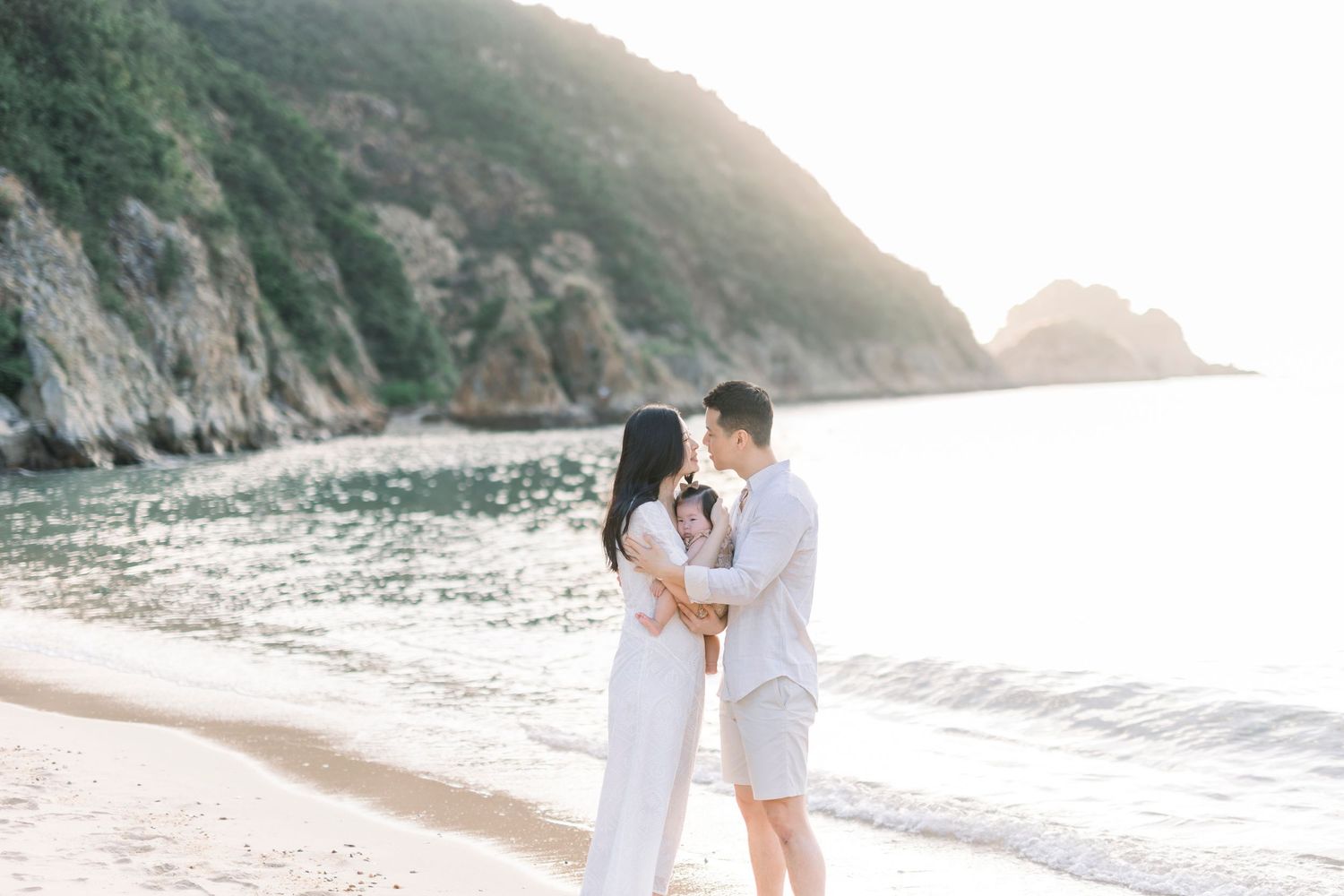 Couple walking together along a serene beach with rocky cliffs and ocean waves in the background.