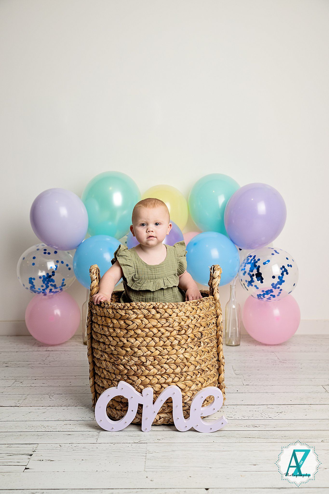 First birthday setup with colorful balloons, woven basket, and 'one' sign on white wooden floor.