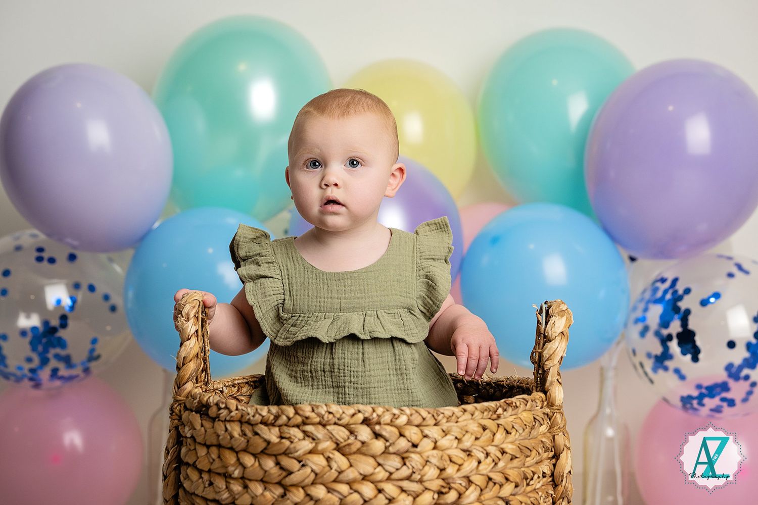 A baby sits in a woven basket surrounded by pastel balloons and confetti against a neutral backdrop.