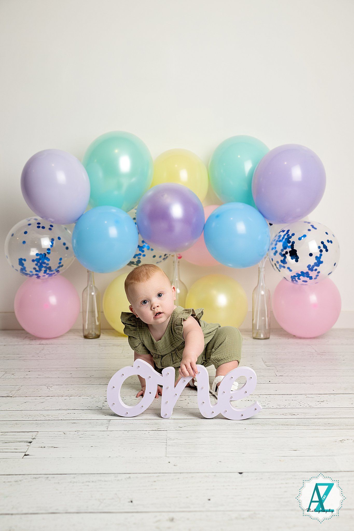 Baby crawls on white wooden floor with pastel colored balloons and letter decorations in studio setting.