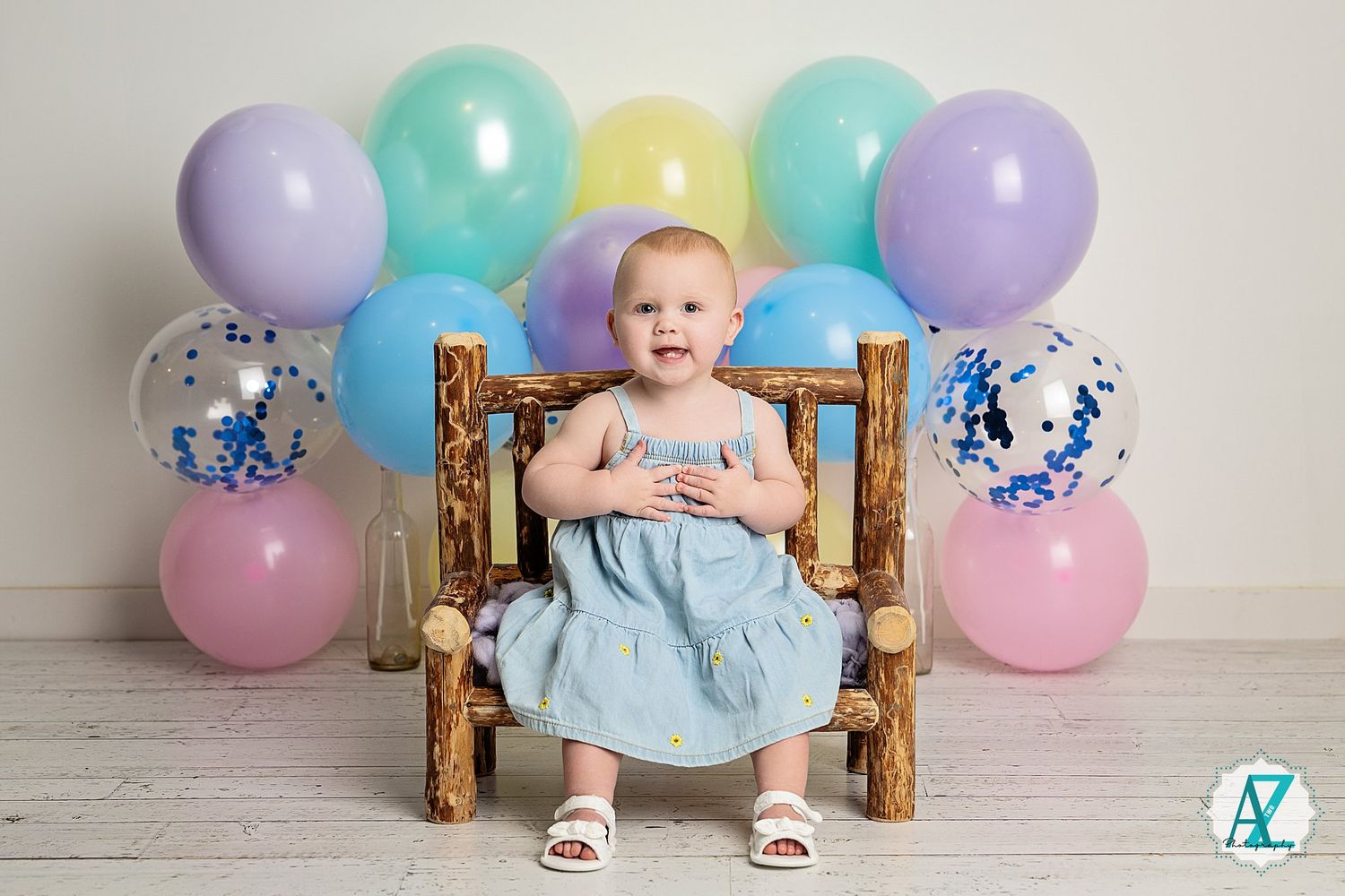 Birthday photo setup with pastel balloons and rustic wooden chair on white floor.