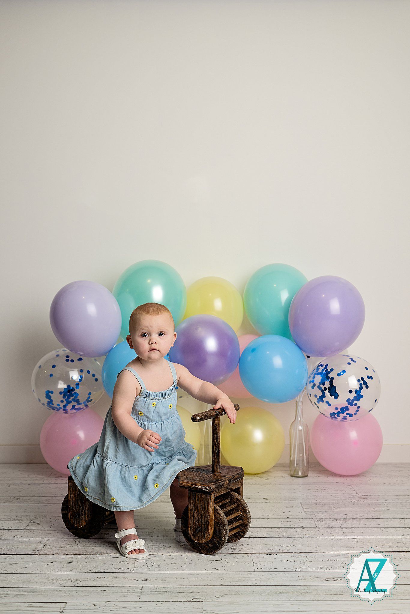 Baby in light blue dress sits against white backdrop with colorful pastel balloons and vintage suitcase.