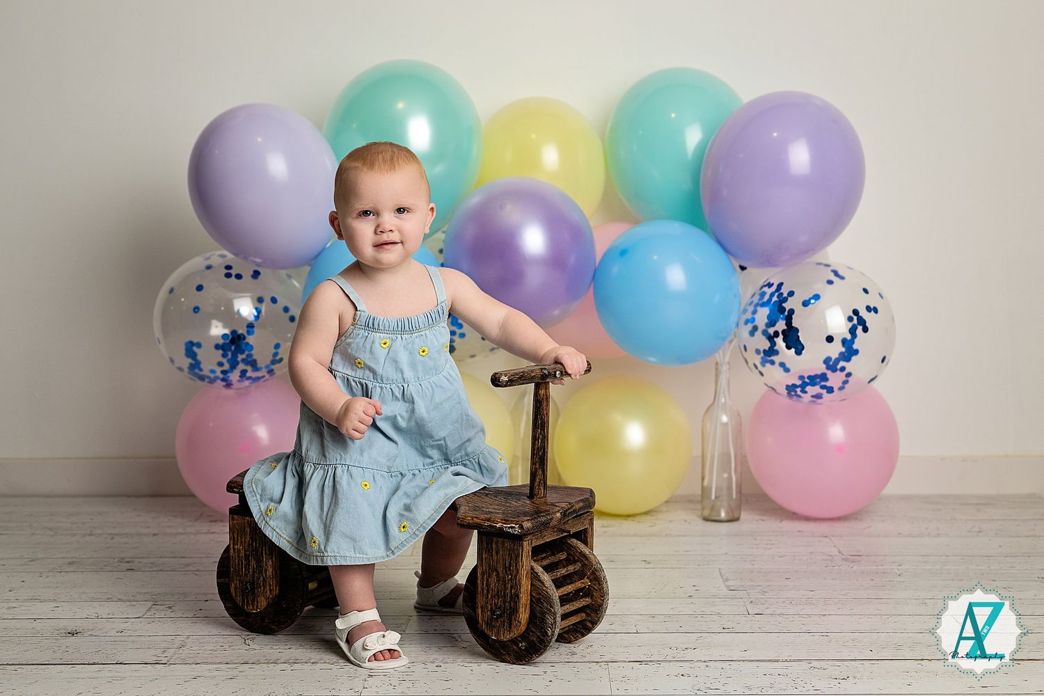 Baby in blue dress sits on wooden floor surrounded by pastel balloons and colorful confetti decorations.