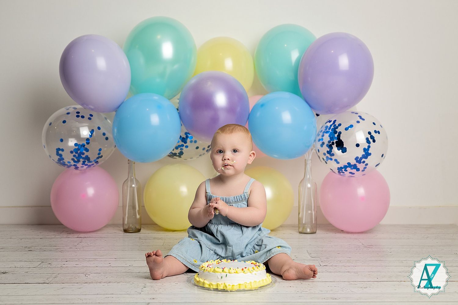 First birthday setup with cake, pastel balloons, and baby sitting on wooden floor against white backdrop.