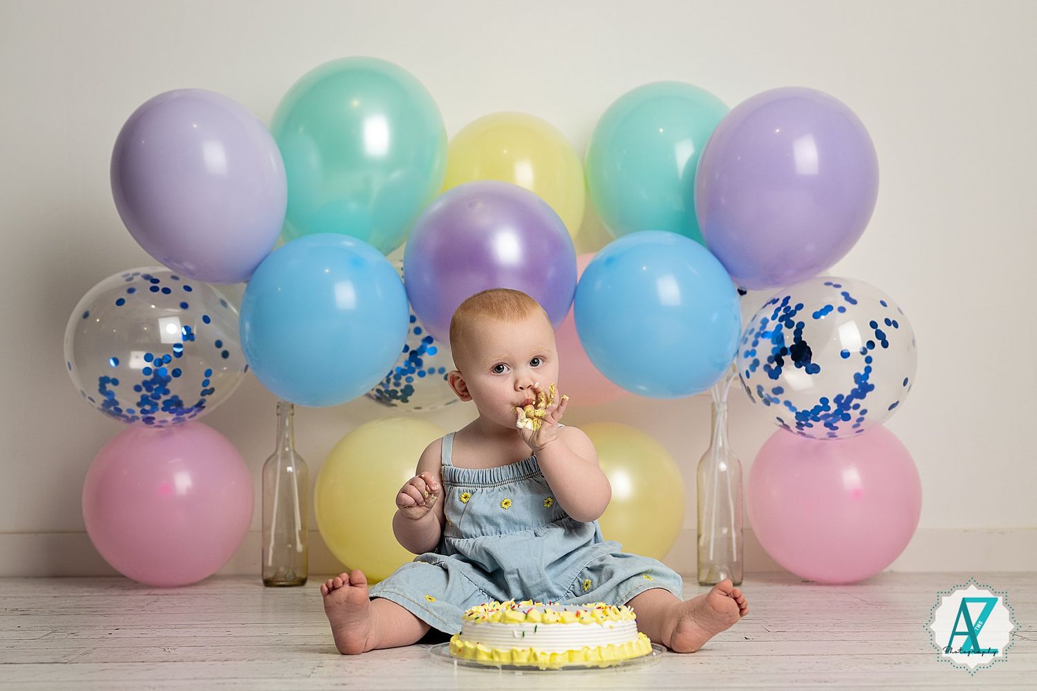 Birthday setup featuring a cake and colorful balloons in mint, lavender, and pink arranged against a light background.