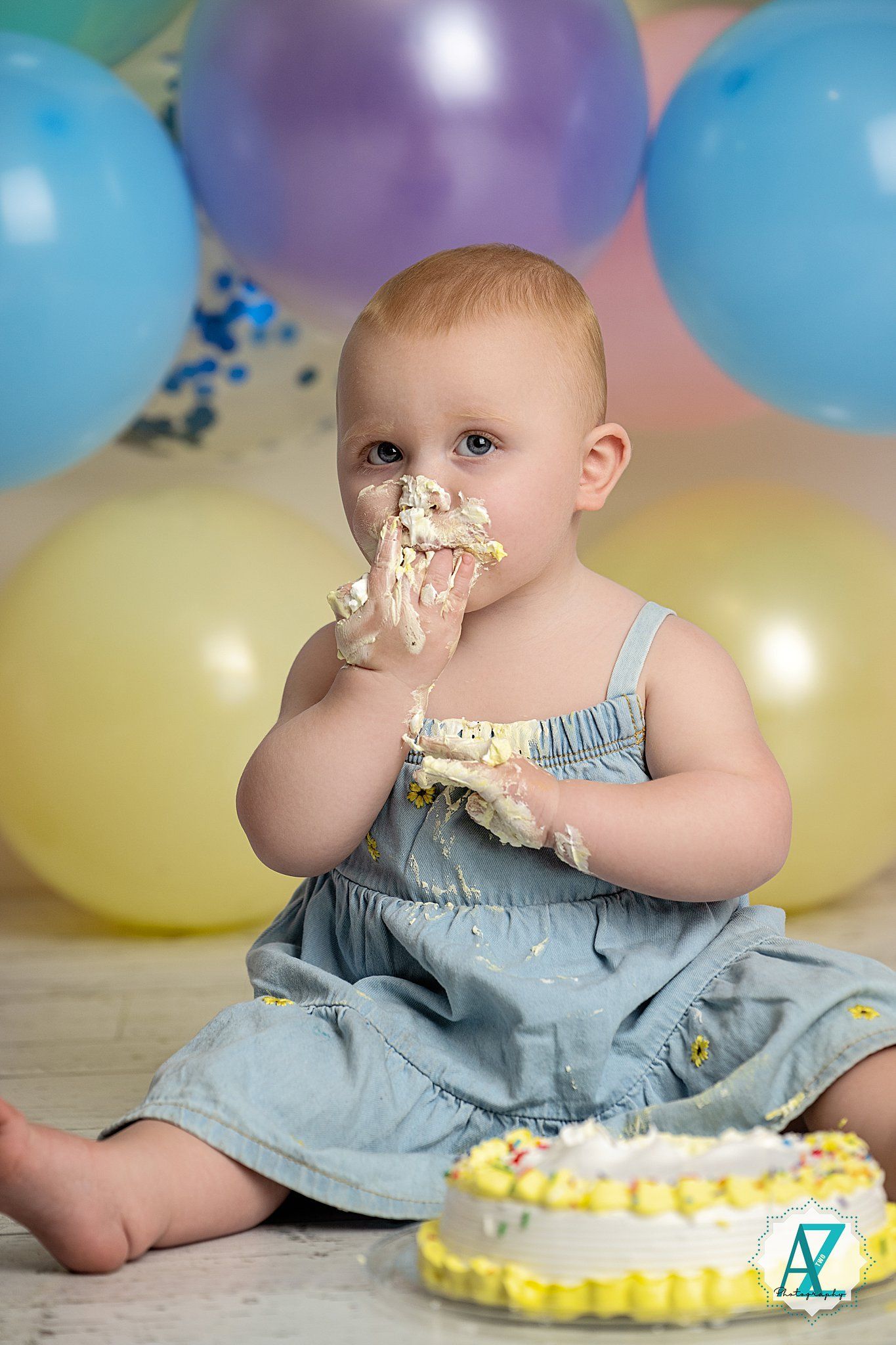Baby in blue dress enjoying cake smash photo session with colorful balloons in background.