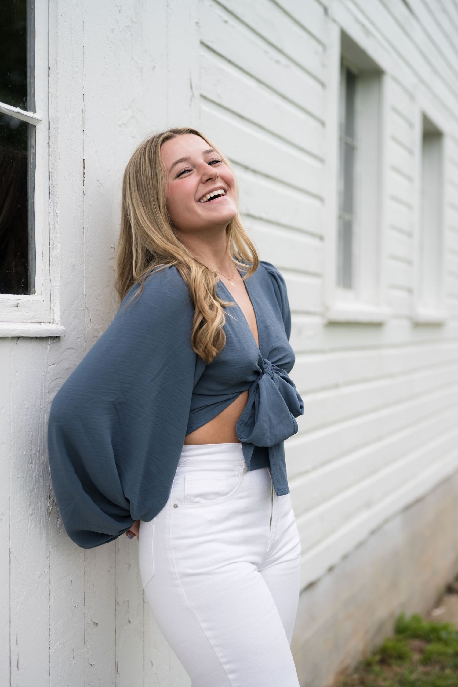 Laughing blonde teenage girl in blue crop top and white jeans leaning against a white building.