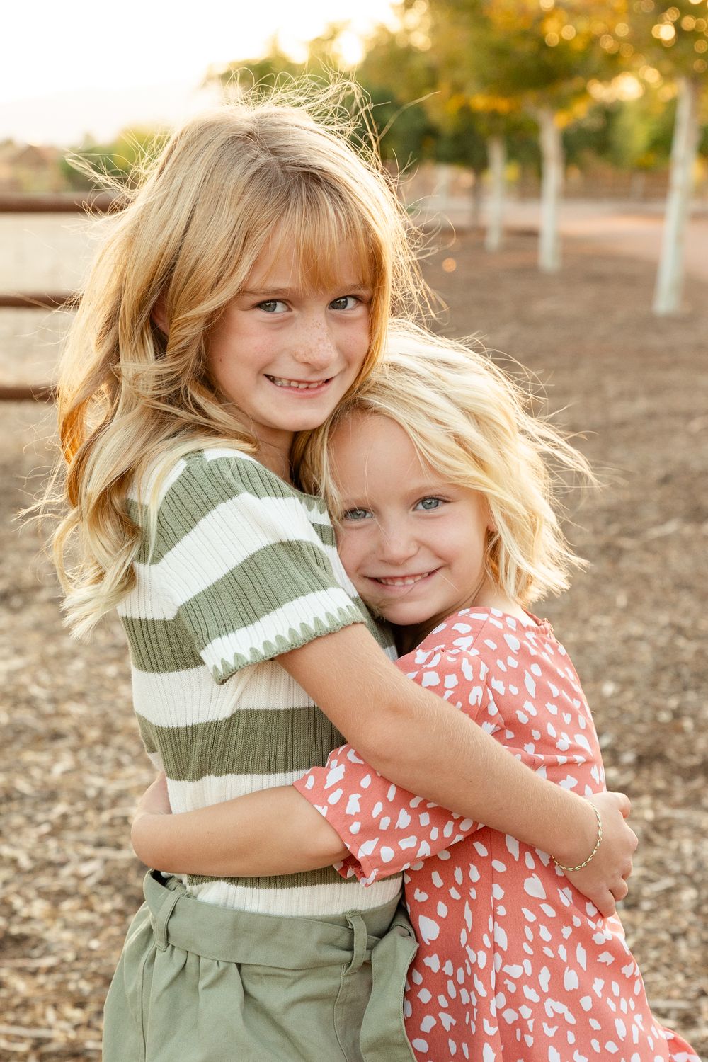 two girls hugging smiling at the camera one is 8 and the other is 5. sisters. farm in the background