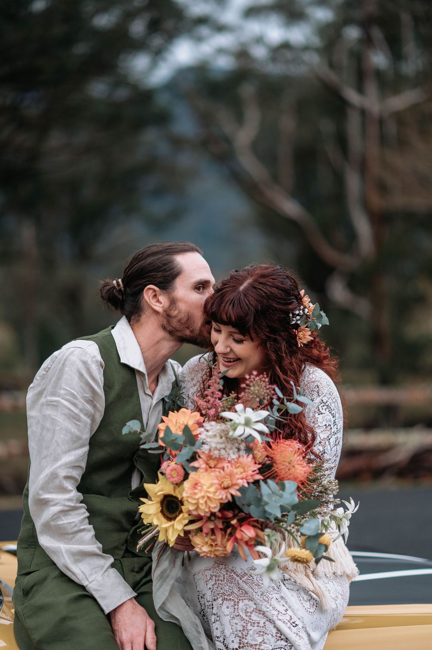 A romantic couple shares tender moments in nature while holding a vibrant autumn-colored wedding bouquet with sunflowers.