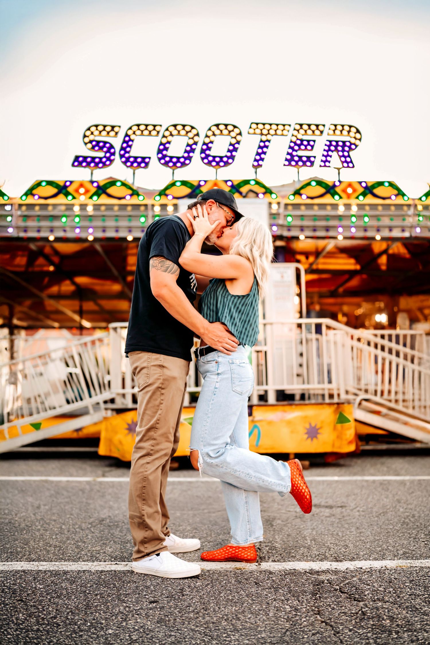 A couple shares a romantic kiss at a carnival with a colorful scooter ride sign illuminated in the background.