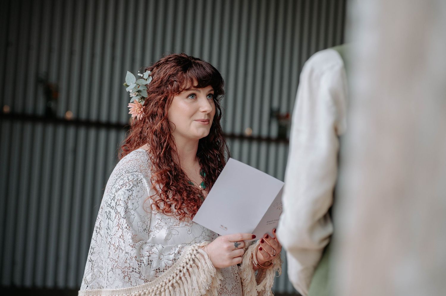 A person wearing a white lace dress holds papers while standing next to a sculpture in an artistic setting.