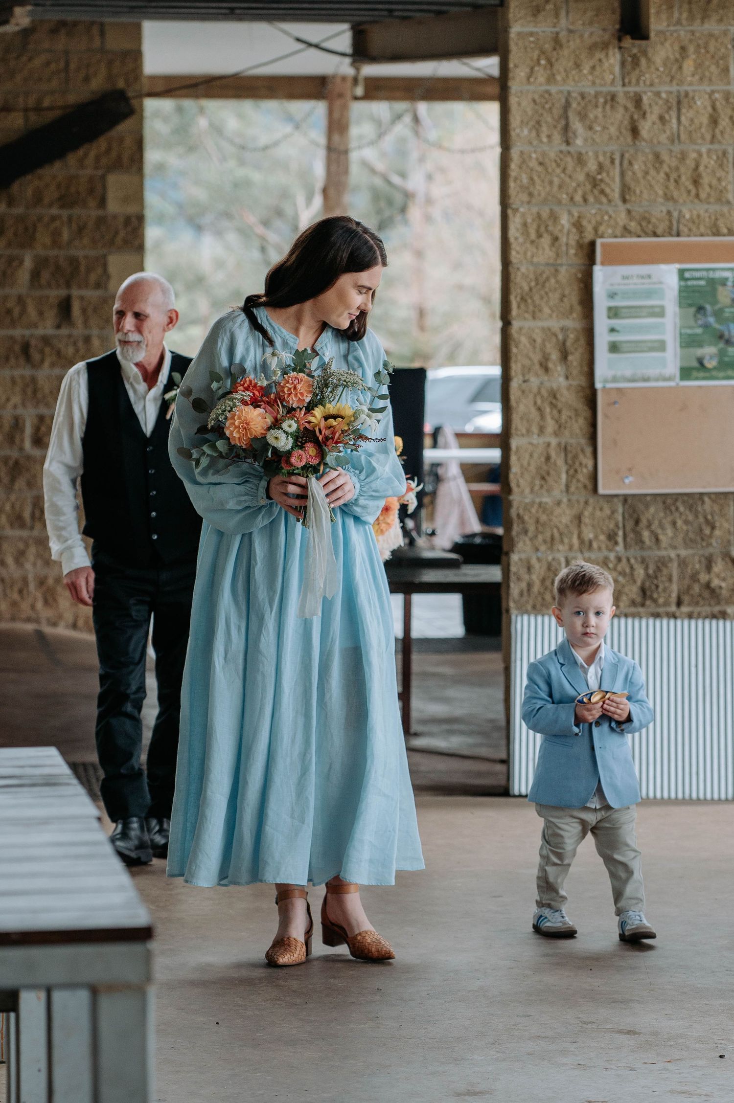 Wedding party members in coordinating light blue outfits gather together holding bouquets in a stone building.