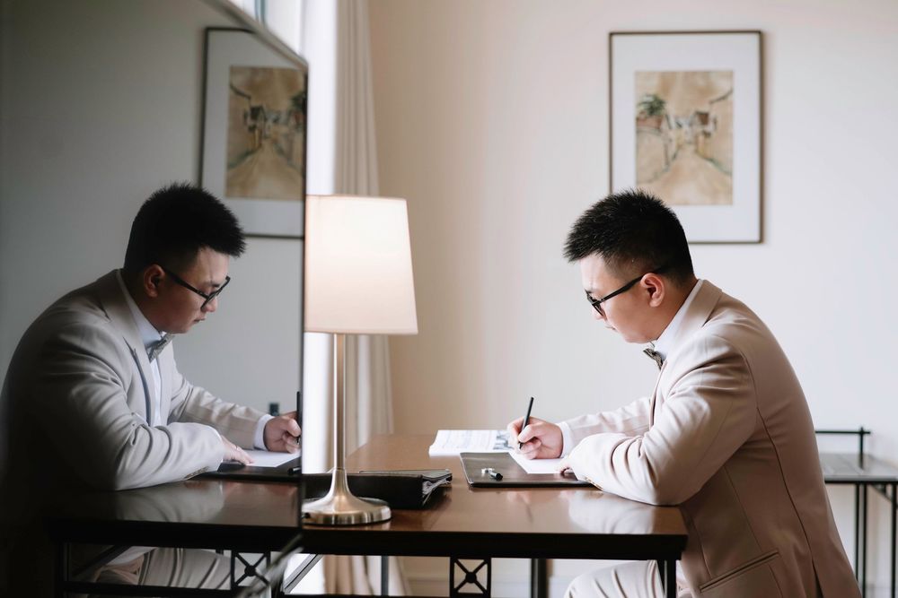 Two men in beige suits sitting across a table writing documents by a lamp.