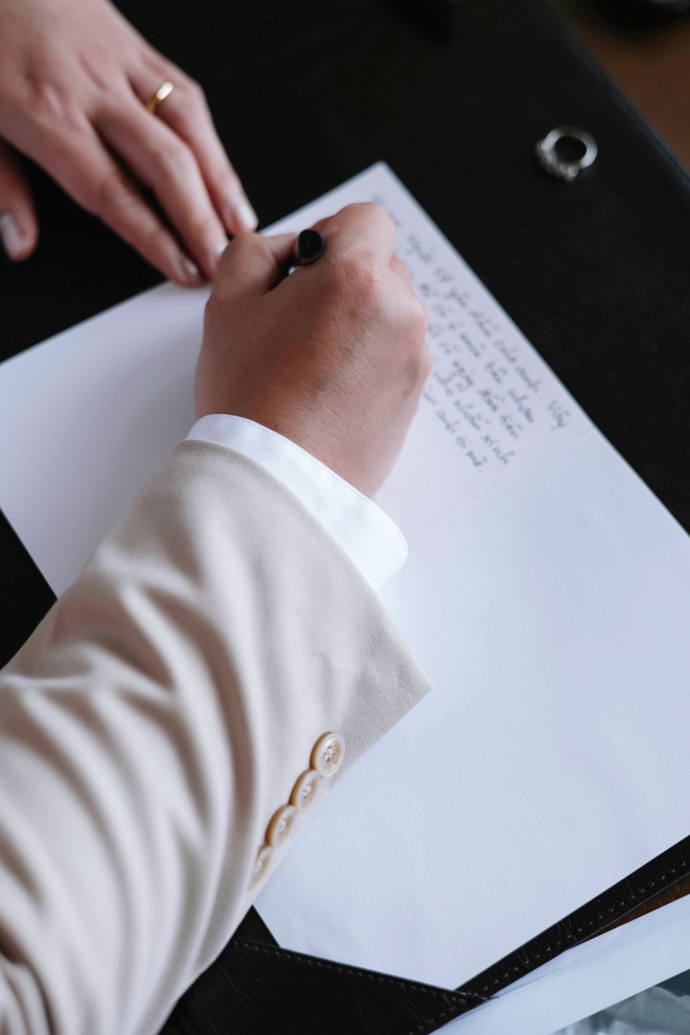 Close-up of groom in beige suit signing wedding documents with a hand resting nearby.