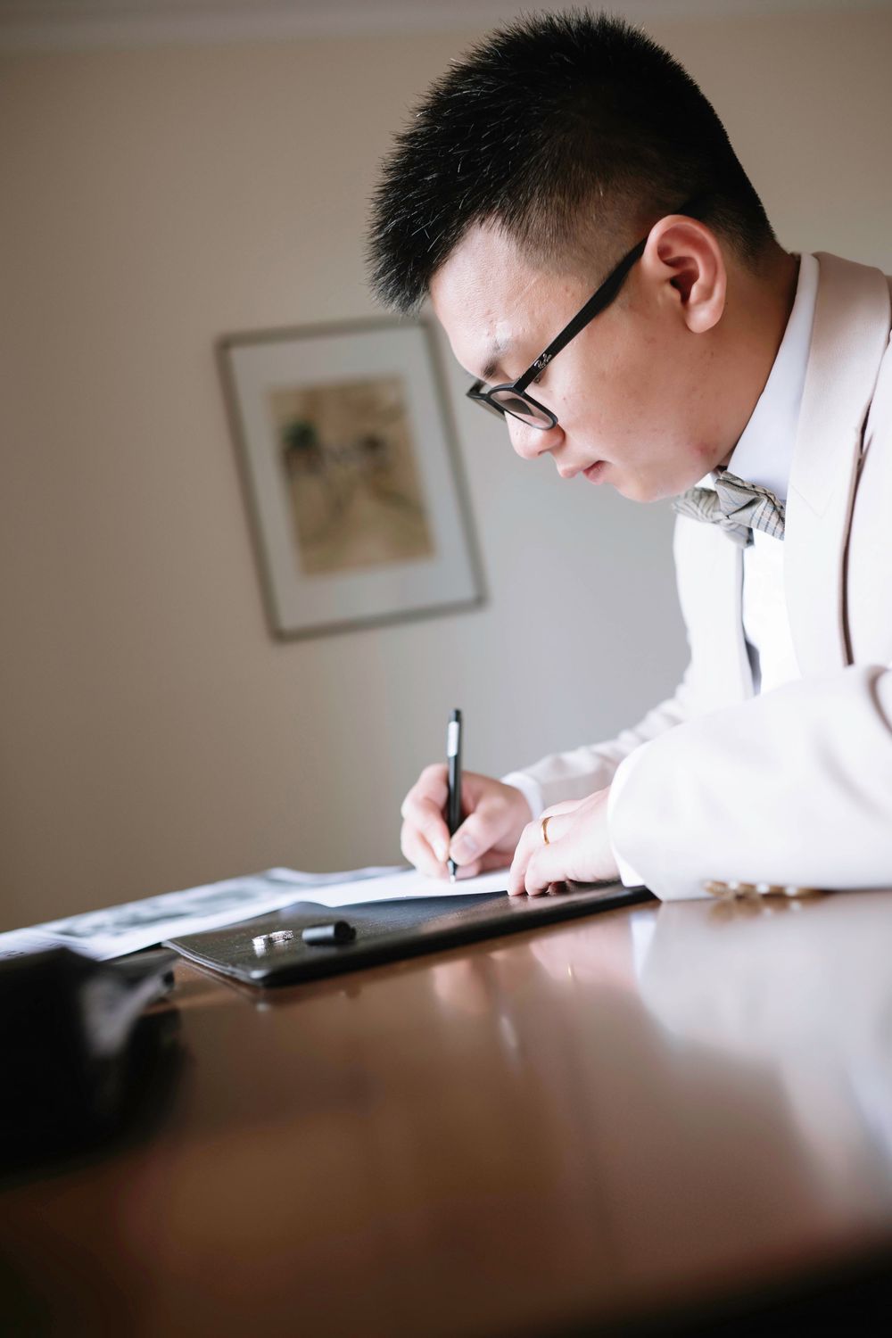Groom in beige suit and bow tie signing documents at a wooden desk.