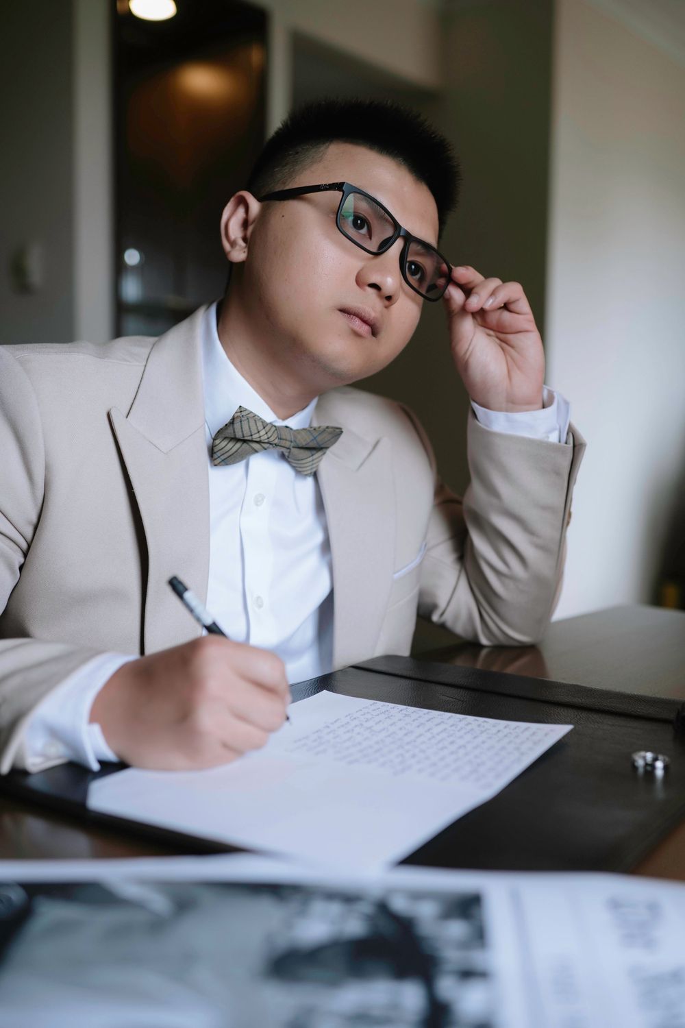 Groom in beige suit adjusting glasses while writing wedding documents at a desk.