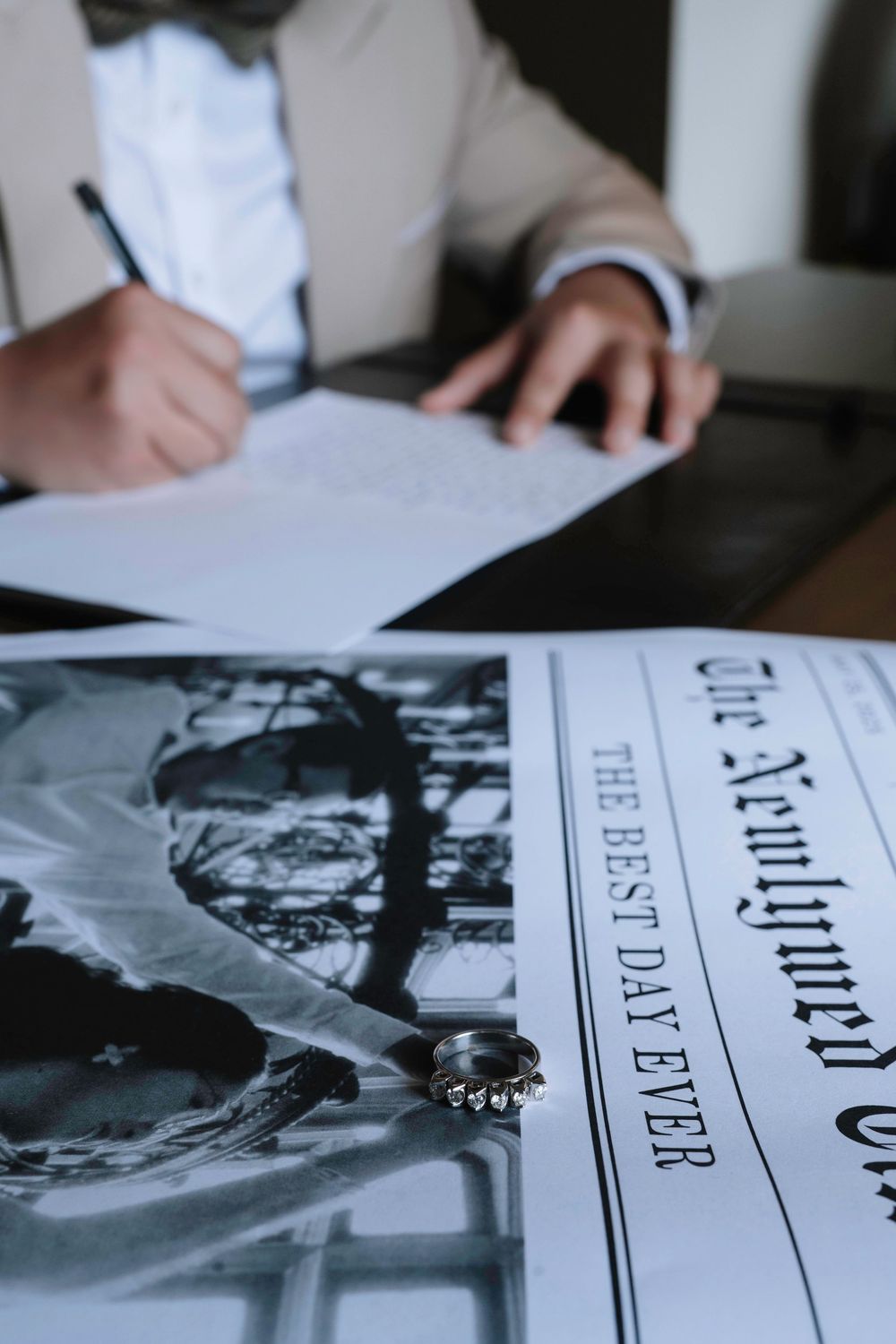 Wedding rings resting on a newspaper beside a groom signing documents at a table.